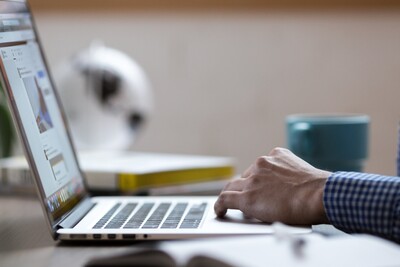 Person in kariertem Hemd arbeitet an einem Laptop, daneben ein Notizbuch und eine Tasse auf einem Tisch.