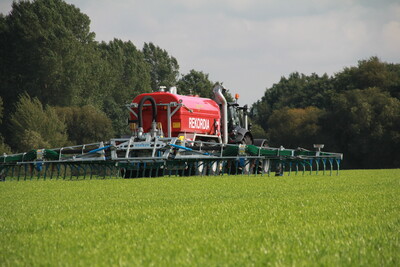 Emissionsarme Gülleausbringung: Schleppschuhverteiler - © Jörn Gläser / Tammo Gläser Productions Landwirtschaftsmaschine mit rotem Tank fährt über ein grünes Feld, im Hintergrund Bäume und bewölkter Himmel.
